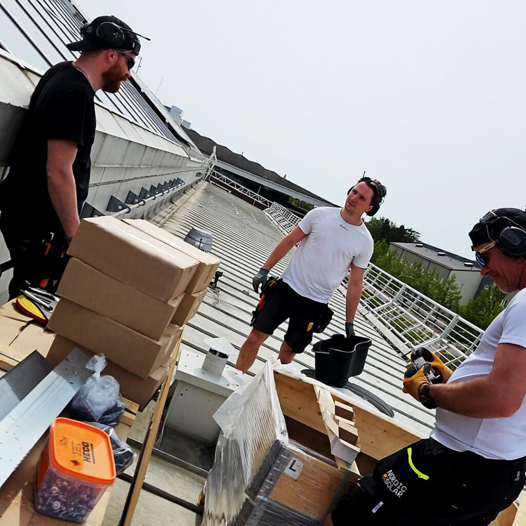 Montörer från Nordic Solar jobbar med att montera solceller på taket till en byggnad under dagtid.