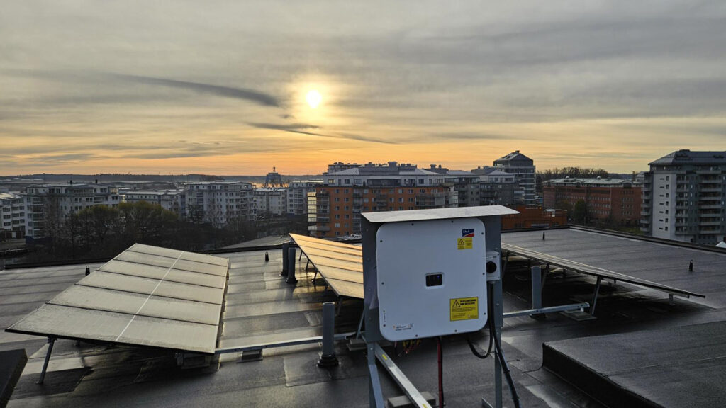 Solcellsanläggning på ett tak i stadsmiljö vid soluppgång, med paneler och växelriktare i förgrunden och flerbostadshus i bakgrunden.